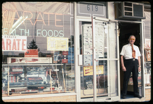 Shop owner standing in the doorway of a store with signs reading 'Karate' and 'Health foods'