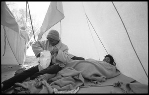 Inside of a strikers' tent: one woman huddled under blankets on the ground, another reaching into a bag