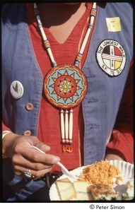 MUSE concert and rally: demonstrator eating food and wearing a Native American beaded necklace and 'The Longest Walk' patch