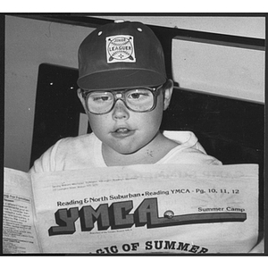 Boy wearing glasses and a baseball cap, looking at a Young Men's Christian Association newsletter