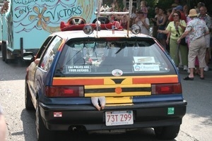Car in the parade with loud speakers, bumper stickers, and a toy doll hanging from the rear hatch : Provincetown Carnival parade