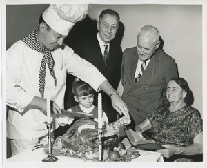 Chef carving turkey with a young client while Mr. Burrows, Mr. Place and an unidentified woman holding a bible look on