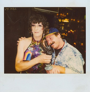 A Photograph of Sylvia Rivera Wearing a Colorful Sequined Dress, Holding a Statuette, and Being Hugged By Another Person