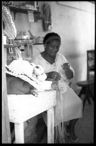 Woman (H. N. Cove) seated by a table in her store, plaiting palm leaf baskets, Belize City