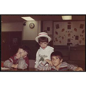 Three boys sit and stand at a table at the South Boston Boys & Girls Club, one wearing a white boater hat and "Southie" hoodie