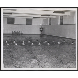 A group of boys swim in a natatorium pool as other boys and a man look on