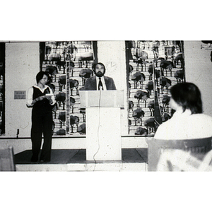 Suzanne Lee stands beside a man who speaks from a podium at a gathering of the Chinese Progressive Association