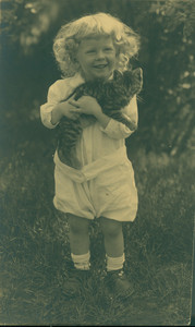 Full-length portrait of a child, standing, facing front, holding a kitten, location unknown, undated