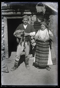 Man, holding a lamb, and woman in front of a hut