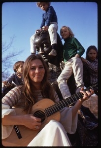 Judy Collins seated at the base of a statue, playing guitar for children