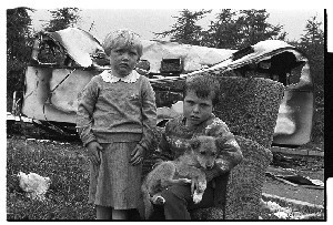 Two children sitting in an old armchair with their dog in front of a burnt-out caravan (their home, allegedly burnt by a fire that was started maliciously by local area residents), Flying Horse Road, Downpatrick