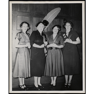 Four women of the Mother's Club pose in front of a feather emblem mounted on the wall