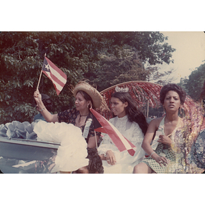 Three women sit on the back of a car during a parade for the Festival Puertorriqueño