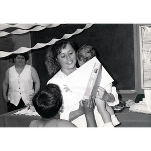 Woman holding a baby, a certificate, and a boxed puzzle during a child care training program and celebration.