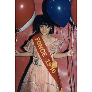 A young girl representing Ponce wears a sash and holds balloons at the Festival Puertorriqueño
