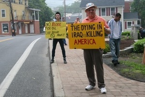 Protesters on the sidewalk holding signs against the War in Iraq reading 'The dying in Iraq is killing America' and 'No blood for oil -- life before profit'