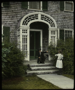 Woman, child, and black cat at the arched entryway to a shingled house