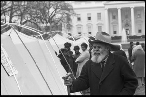 Bearded man with cowboy hat and cane walks past a tent, the White House in the background