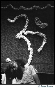 Ram Dass seated on stage, eyes shut, touching foreheads with an unidentified man during his appearance at President Theodore Roosevelt High School