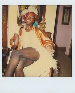 A Photograph of Marsha P. Johnson Sitting With Her Legs Crossed, Smiling Wearing a Tulle Headpiece and in a White Orange Dress