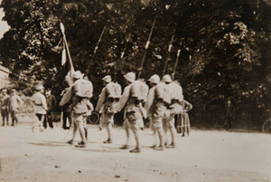 View of soldiers with bayonets marching along a tree-lined street as civilians look on