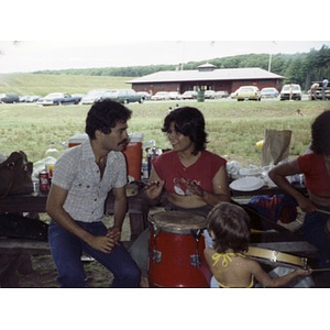 Smiling woman is seated at a picnic table playing the drums at a La Alianza staff picnic, while a man sits next to her.