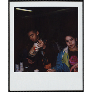 A boy holding a root beer bottle while a girl stands beside him and looks off during an event at the Chelsea Clubhouse