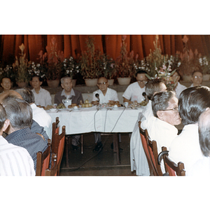 Men sit at banquet tables in front of a stage decorated with potted plants