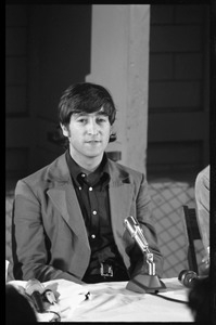 John Lennon seated in front of a microphone at a table, during a Beatles press conference