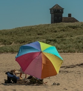 Woman with an umbrella pitched on the beach, with the life saving station in the background, Provincetown