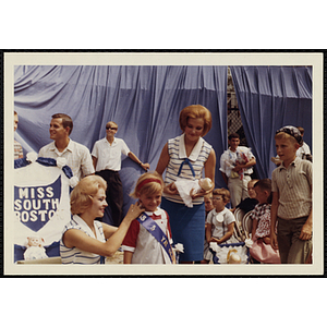 Two women present a sash and a baby doll to Miss 5 Year Old while her brother looks on during a Little Sister Contest