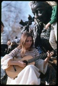 Judy Collins seated at the base of a statue, playing guitar for children