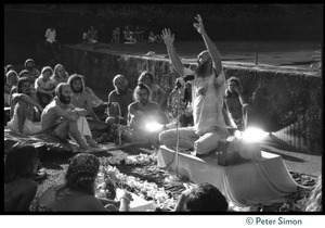 Ram Dass seated on a small platform, arms raised, during his appearance at Andrews Amphitheater, University of Hawaii