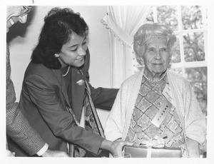 Deirdre A. Ling standing indoors, handing gift to Margarita Hopkins Rand