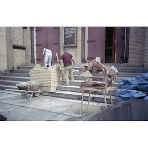 Two workmen building a brick divider on the front steps of the Jorge Hernandez Cultural Center.