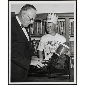 The Boys' Club Freckle King Contest winner holding his prize and looking at a man standing at the typewriter