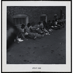 A dozen of boys sit against a wall and eat at a Boys' Club Little Sister Contest