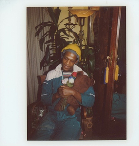 A Photograph of Marsha P. Johnson Wearing a Construction Hat and Tracksuit, Holding a Stuffed Bear and Flowers