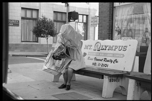 Woman with load of dry cleaning, seated on a bench on a Los Angeles Street