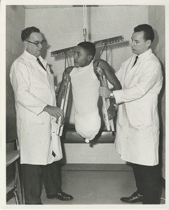 Two technicians help a double amputee into a pair of hanging crutches to aid in a prosthetic fitting