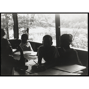 Four boys sit at a table with pencils, paper and scissors