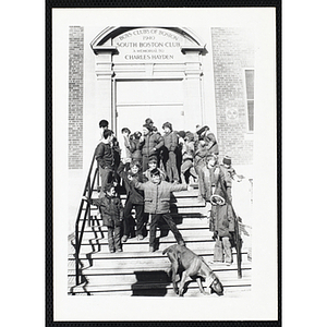 About fifteen Boys & Girls Club members stand on the front steps of the South Boston Clubhouse while a dog goes down the steps in the foreground