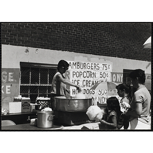 Two girls and a woman watch a woman make cotton candy at a carnival