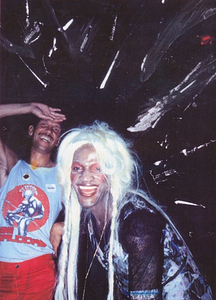 A Photograph of Marsha P. Johnson Leaning Forward and Laughing with Blonde Hair and a Black and Pink Patterned Dress