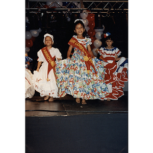 Young girls on stage wearing dresses and sashes at the Festival Puertorriqueño