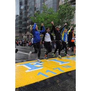 Five runners, holding hands with arms raised, begin crossing the "One Run" finish line at Copley Square