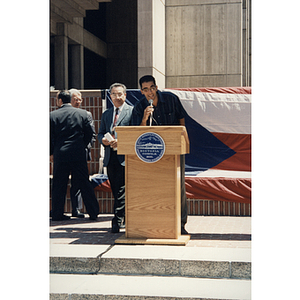 A man speaks at a lectern while another man stands beside him