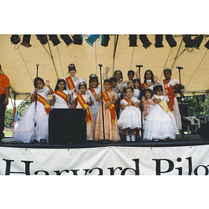 Several women and young girls stand on stage wearing dresses and sashes at the Festival Puertorriqueño