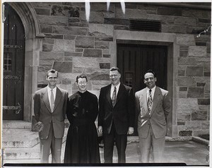 Four men standing outside building at Boston College, one is a member of the clergy