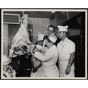 A member of the Tom Pappas Chefs' Club cuts into meat on a hook in a Brandeis University kitchen as two fellow members, a kitchen staff member, and Chairman of Chefs Club Committee Alexander Armour (wearing glasses) look on